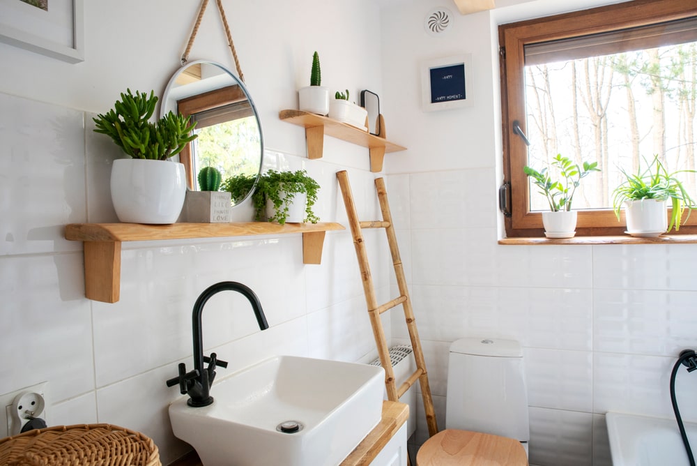 Interior of a small bathroom with a window, wooden decor, shelves, and a ladder. Features an elegant sink and faucet, a round mirror on a white wall, and plants on a shelf.