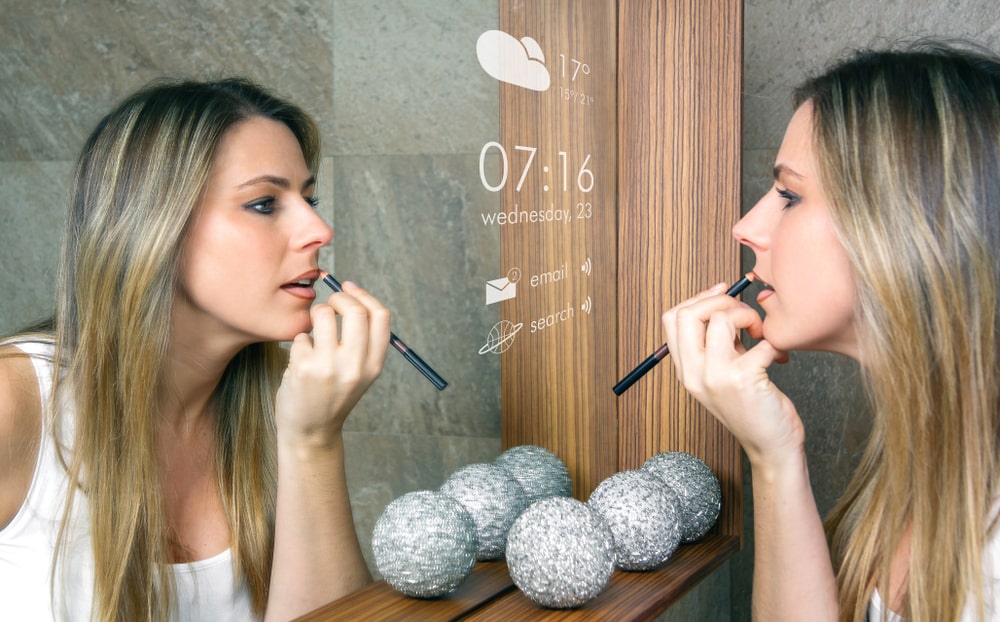 A young woman applying a lip balm in front of a smart mirror that provides information.